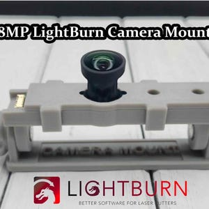 May include: A gray 3D printed camera mount for an 8MP camera. The mount has a slot for the camera lens and two holes on either side. The camera is mounted on the mount and is pointing towards the right. The text "CAMERA MOUNT" is printed on the front of the mount. The text "LightBurn" is printed on the bottom of the image.