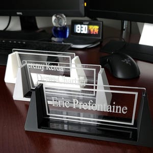 May include: Three clear acrylic desk nameplates with white lettering on black stands. The nameplates read "Jeremy Korda", "Denis Chartier", and "Eric Prefontaine".