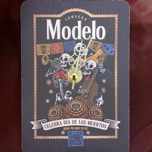 Puede incluir: Un reloj negro y dorado con una etiqueta de cerveza Modelo. El reloj presenta un tema del Día de Muertos con esqueletos tocando instrumentos musicales. El texto "Celebra Dia de los Muertos" y "Bring the Band to Life" está impreso en el reloj.