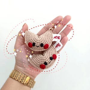 May include: Two crocheted fortune cookie charms with red cheeks and black eyes. The charms are beige and have the word "ATE" written on them in red.