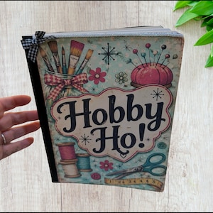May include: A rectangular book with a light blue cover featuring illustrations of paintbrushes in a jar, a pink pincushion, spools of thread, scissors, and a measuring tape. The text Hobby Ho! is printed in large black letters.