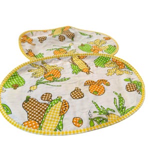 May include: Two oval-shaped fabric placemats featuring a white background with a repeating pattern of vegetables including corn, onions, and peppers in shades of orange, green, and brown, finished with a yellow and white checkered fabric trim.