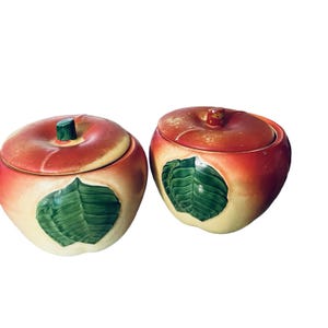 May include: Two ceramic apple-shaped lidded containers featuring a gradient of red and cream colors. Each container has a sculpted green leaf on the front and a small green stem on the lid. The finish shows signs of wear with some paint loss.