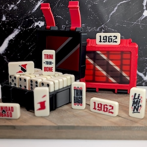 May include: A set of off-white dominoes with black pips and red accents, featuring text including 1962, Trini To Di Bone, Trinbago, and Limin. The set is housed in a black and red carrying case with the Trinidad and Tobago flag design.