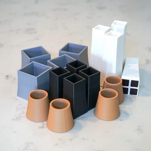 May include: An assortment of geometric, minimalist decor pieces in various shapes and colors. Includes square and rectangular vases in light blue, black, and white, along with several terracotta-colored, cone-shaped objects. The objects are arranged on a white surface.