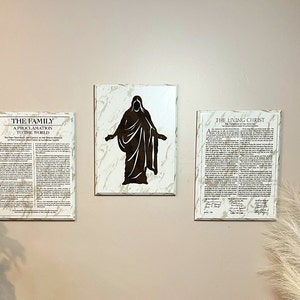 Puede incluir: Tres impresiones enmarcadas en una pared de color beige claro. La impresión de la izquierda es una proclamación sobre la familia. La impresión del medio es una silueta de una persona con los brazos extendidos sobre un fondo de mármol blanco. La impresión de la derecha es una proclamación sobre el Cristo viviente.