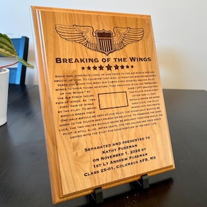 Peut inclure: Une plaque en bois avec une image gravée en brun d'une paire d'ailes avec une étoile au centre. Le texte "Breaking of the Wings" est gravé en police brune avec cinq étoiles sous le texte. La plaque est séparée et présentée à Kathy Puseman le 1er novembre 2024 par le 1er lieutenant Andrew Puseman, classe 25-01, Columbus AFB, MS.