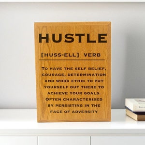 May include: A rectangular wooden plaque with the word "HUSTLE" in bold black letters. Below, the definition of "Hustle" is printed in black text. The plaque is made of light brown wood and is displayed on a white surface.