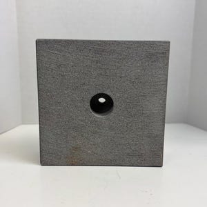 May include: A gray, square block with a circular hole in the center. The block has a textured surface and appears to be made of stone or a similar material. The hole is centered on one of the block's faces.