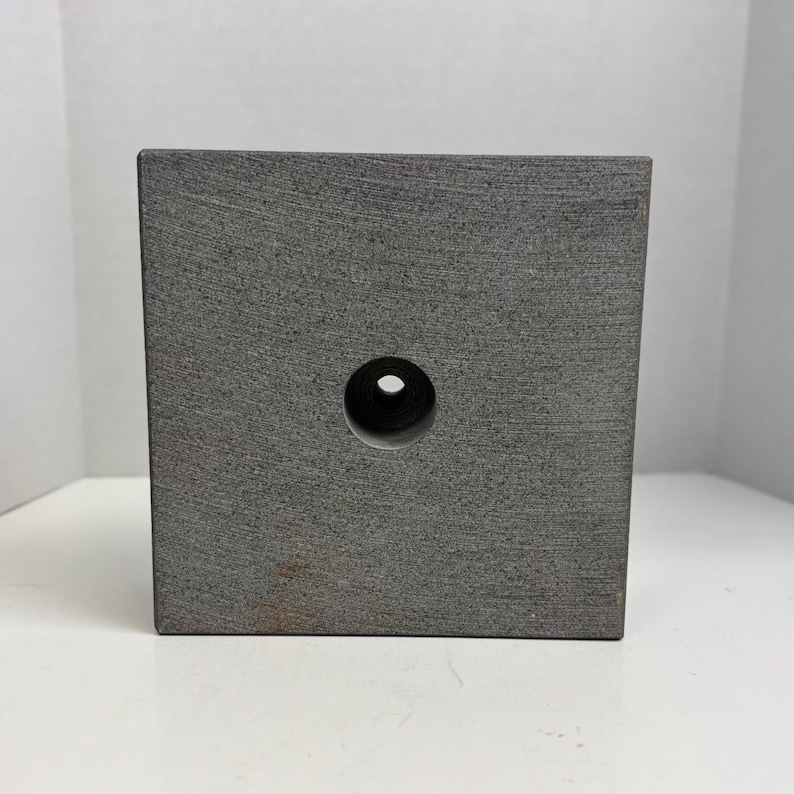 May include: A gray, square block with a circular hole in the center. The block has a textured surface and appears to be made of stone or a similar material. The hole is centered on one of the block's faces.