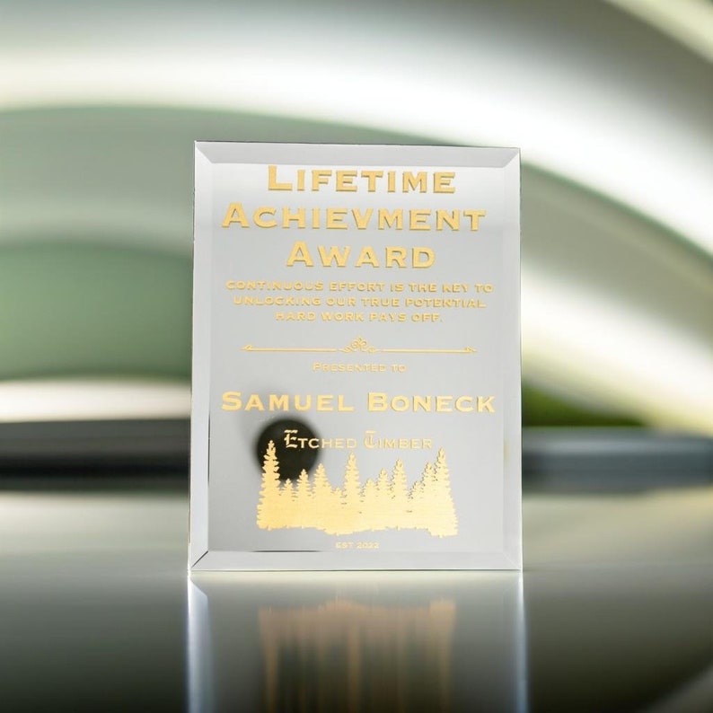 Puede incluir: Un premio de vidrio transparente con letras doradas. El texto dice "LIFETIME ACHIEVEMENT AWARD" y "SAMUEL BONECK". Debajo del nombre est&aacute; "ETCHED TIMBER" y un gr&aacute;fico de bosque. El premio es rectangular y est&aacute; en posici&oacute;n vertical.
