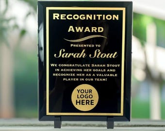 May include: Black plaque with gold lettering recognizing Sarah Stout for achieving her goals. The plaque reads "Recognition Award" and "We congratulate Sarah Stout in achieving her goals and recognize her as a valuable player in our team!"  The plaque has a gold circle with the text "Your Logo Here".