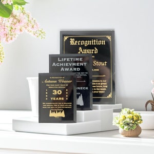 Puede incluir: Una colecci&oacute;n de premios de reconocimiento en negro y dorado. Los premios incluyen textos como "Premio de Reconocimiento", "Premio a la Trayectoria" y un premio personalizado para Autumn Weaver, que celebra 30 a&ntilde;os de servicio. Los premios se exhiben en plataformas blancas.