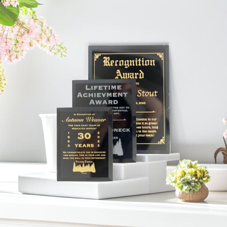 Puede incluir: Una colecci&oacute;n de premios de reconocimiento en negro y dorado. Los premios incluyen textos como "Premio de Reconocimiento", "Premio a la Trayectoria" y un premio personalizado para Autumn Weaver, que celebra 30 a&ntilde;os de servicio. Los premios se exhiben en plataformas blancas.