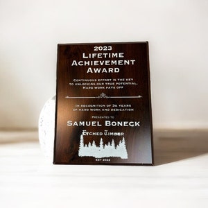 Pu&ograve; includere: Una targa in legno marrone con testo bianco che recita "2023 Lifetime Achievement Award". La targa ha una silhouette bianca di una foresta e il testo "Etched Timber Est 2022".