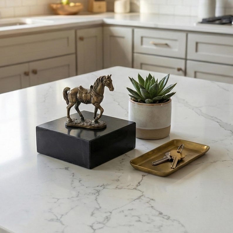 May include: A bronze horse statue on a black square base, a small succulent in a beige and white pot, and a gold-colored rectangular tray with keys are displayed on a white countertop. The horse statue is detailed with a saddle.