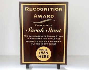 May include: A dark brown wooden plaque with gold lettering. The plaque reads "Recognition Award" and "Presented to Sarah Stout". The plaque also includes the text "We congratulate Sarah Stout in achieving her goals and recognize her as a valuable player in our team!" and a circle with the text "Your Logo Here".