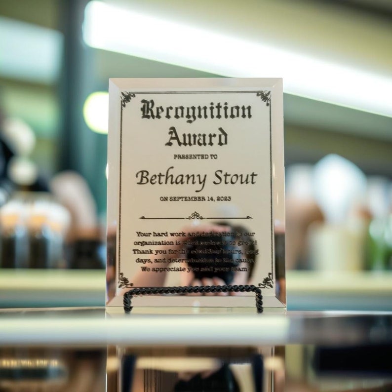Puede incluir: Un premio de reconocimiento de vidrio transparente con texto negro. El premio dice "Recognition Award" y se presenta a "Bethany Stout" el 14 de septiembre de 2023. El premio se muestra en un soporte de metal negro.