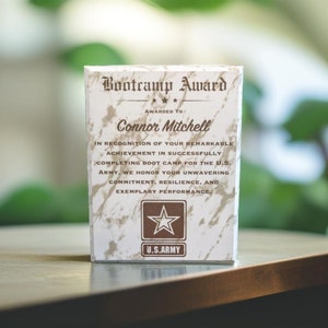 May include: A white plaque with a brown marble pattern and gold lettering. The plaque reads "Bootcamp Award" and "Awarded to Connor Mitchell". The plaque also includes the text "In recognition of your remarkable achievement in successfully completing boot camp for the U.S. Army, we honor your unwavering commitment, resilience, and exemplary performance." The plaque features a gold star and the words "U.S. Army".