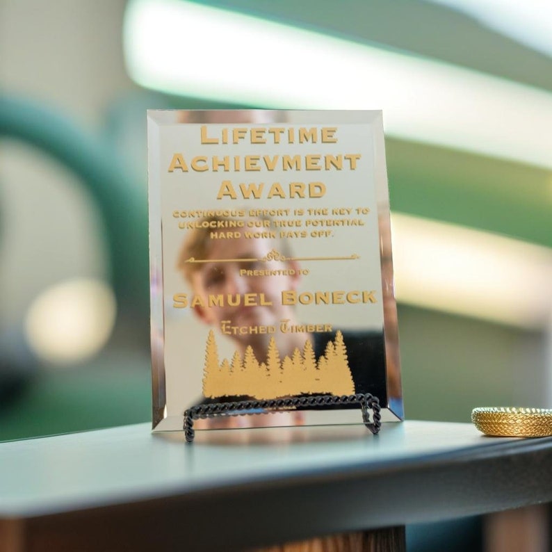 Puede incluir: Un premio de vidrio rectangular con letras doradas que dice "LIFETIME ACHIEVEMENT AWARD". Tambi&eacute;n es visible el texto "Presented to Samuel Boneck". El premio se exhibe en un soporte de metal negro.