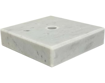 May include: A square, white marble base with a centered hole. The marble has subtle gray veining and a polished finish. The base is a solid, rectangular shape, suitable for display or support.