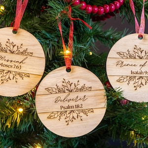 Puede incluir: Tres adornos redondos de madera con un acabado rústico y un diseño floral. Cada adorno tiene una inscripción diferente: "Mesías Moisés 7:63", "Libertador Salmo 18:2" y "Príncipe de Paz Isaías 9:6".
