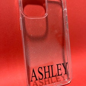Op de afbeelding: Transparante telefoonhoes met een glinsterende afwerking en de naam "ASHLEY" in zwarte letters.