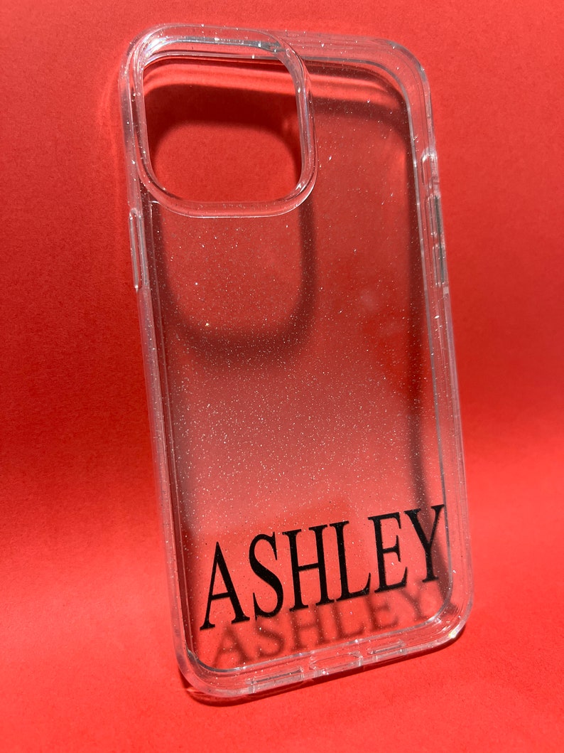 Op de afbeelding: Transparante telefoonhoes met een glinsterende afwerking en de naam "ASHLEY" in zwarte letters.