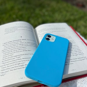May include: A light blue silicone phone case for an iPhone 13 Pro. The case is sitting on top of an open book with the text "I was some distance away, my arms full..." visible.