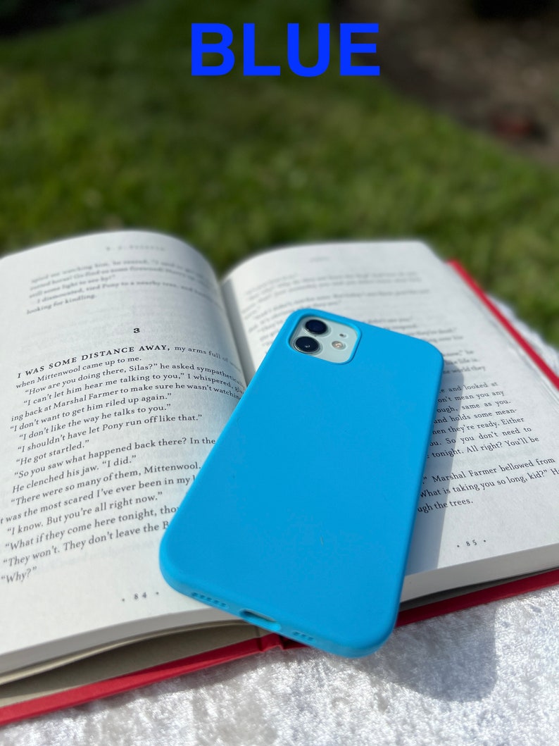 May include: A light blue silicone phone case for an iPhone 13 Pro. The case is sitting on top of an open book with the text "I was some distance away, my arms full..." visible.
