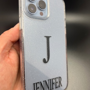 Op de afbeelding: Een transparante telefoonhoes met een lichtblauwe glitterachtergrond. De hoes heeft een zwarte letter "J" en de naam "JENNIFER" erop gedrukt.