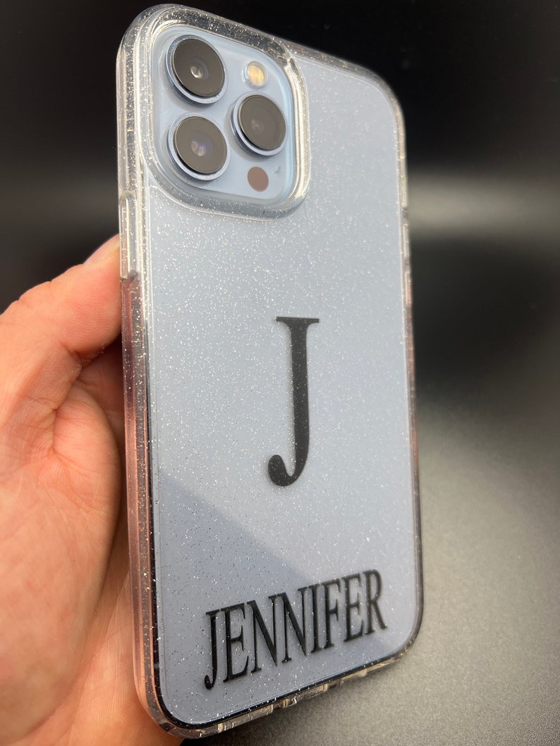 Op de afbeelding: Een transparante telefoonhoes met een lichtblauwe glitterachtergrond. De hoes heeft een zwarte letter "J" en de naam "JENNIFER" erop gedrukt.