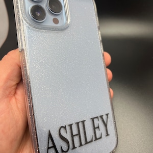Op de afbeelding: Een transparante telefoonhoes met een glinsterende lichtblauwe achtergrond en de naam "ASHLEY" in zwart gedrukt op de achterkant.