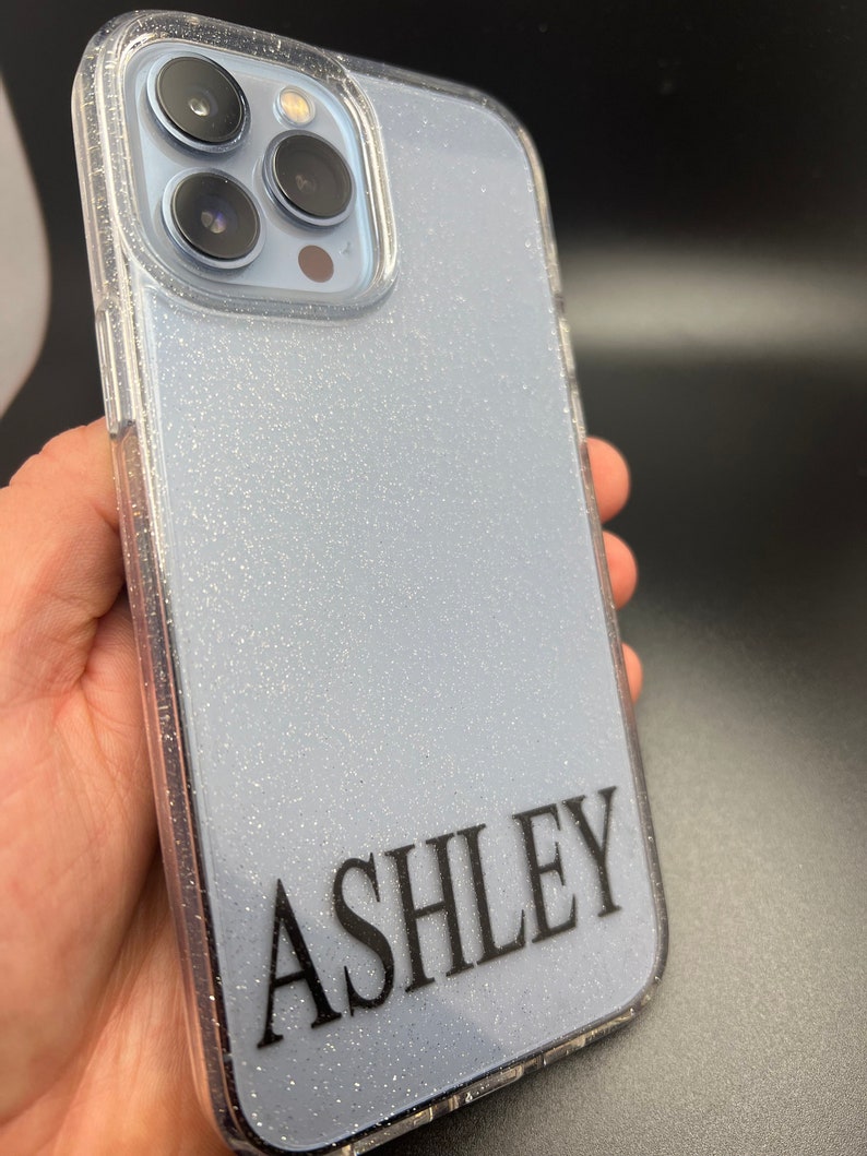 Op de afbeelding: Een transparante telefoonhoes met een glinsterende lichtblauwe achtergrond en de naam "ASHLEY" in zwart gedrukt op de achterkant.