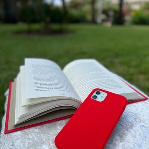 May include: A bright red phone case for an iPhone. The case is made of a soft, flexible material and has a smooth finish. The case is lying on top of a book.