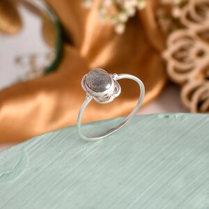 May include: A silver ring featuring an oval, grey gemstone. The gemstone is set in a decorative bezel, and the ring band is thin and simple. The ring is displayed on a light blue surface with a gold and green background.