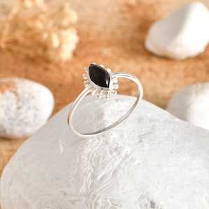May include: A silver ring featuring a marquise-shaped black gemstone. The gemstone is set in a decorative bezel with small, round details. The ring is shown on a white, textured surface, with a blurred background of sand and white stones.