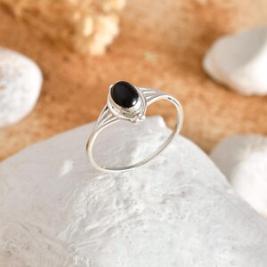 May include: A silver ring featuring an oval, black gemstone. The ring has a slender band and a decorative setting. The ring is shown on a white, textured surface.
