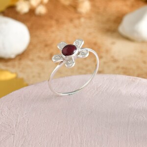 May include: A silver ring with a flower-shaped design. The ring has a dark red central stone and six petals. The ring is set on a thin band. The ring is displayed on a pink surface.