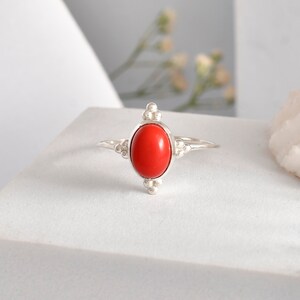 May include: A silver ring with an oval, bright red gemstone. The ring has decorative elements on the sides and top, with a simple band. The gemstone is approximately 1.3 cm in length.