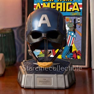 May include: A blue Captain America helmet replica with a large white "A" on the front, displayed on a gray base. The base has the word "INFINITY" on it. A framed Captain America comic book is in the background.