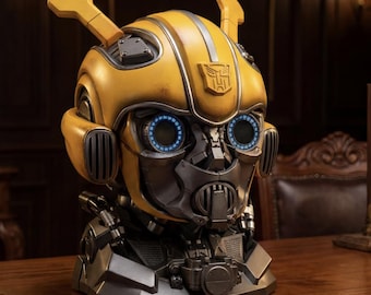 May include: A detailed, yellow Bumblebee head statue with silver accents and blue eye details. The Transformers logo is visible on the forehead. The statue is set on a dark, metallic base, showcasing intricate mechanical features.