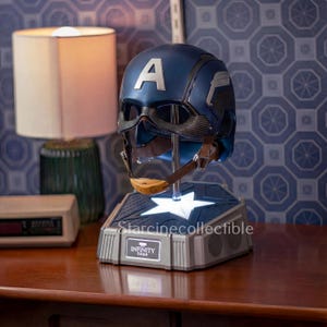 May include: A blue and silver Captain America helmet with the letter "A" on the front, displayed on a light-up base. The base has the text "Starcinecollectible" and "INFINITY SAGA". The helmet has a brown chin strap.