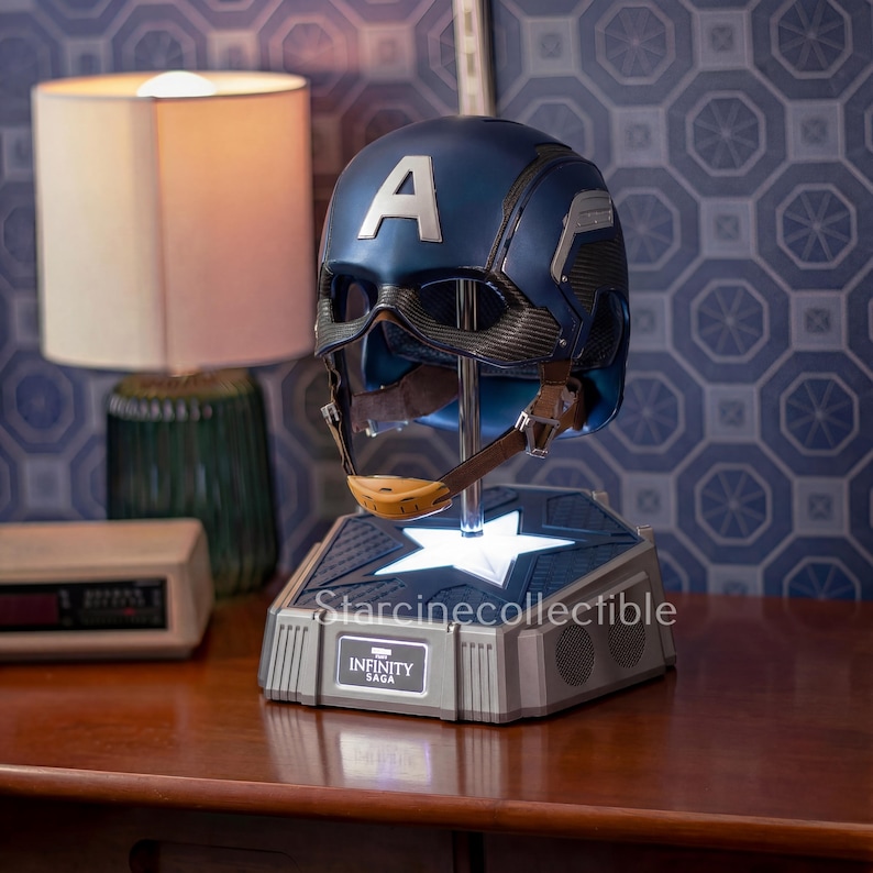May include: A blue and silver Captain America helmet with the letter "A" on the front, displayed on a light-up base. The base has the text "Starcinecollectible" and "INFINITY SAGA". The helmet has a brown chin strap.