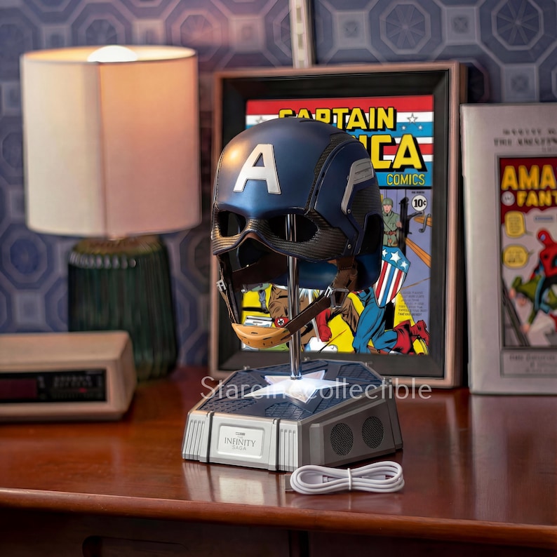 May include: A Captain America helmet replica, featuring a blue and silver design with a prominent white "A" on the forehead. The helmet is displayed on a stand with the word "INFINITY" and a USB cable. A framed comic book is in the background.