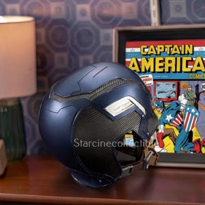 May include: A navy blue Captain America helmet with silver accents and a carbon fiber-style mesh. The helmet is displayed next to a framed Captain America comic book cover. The helmet has a brown strap.