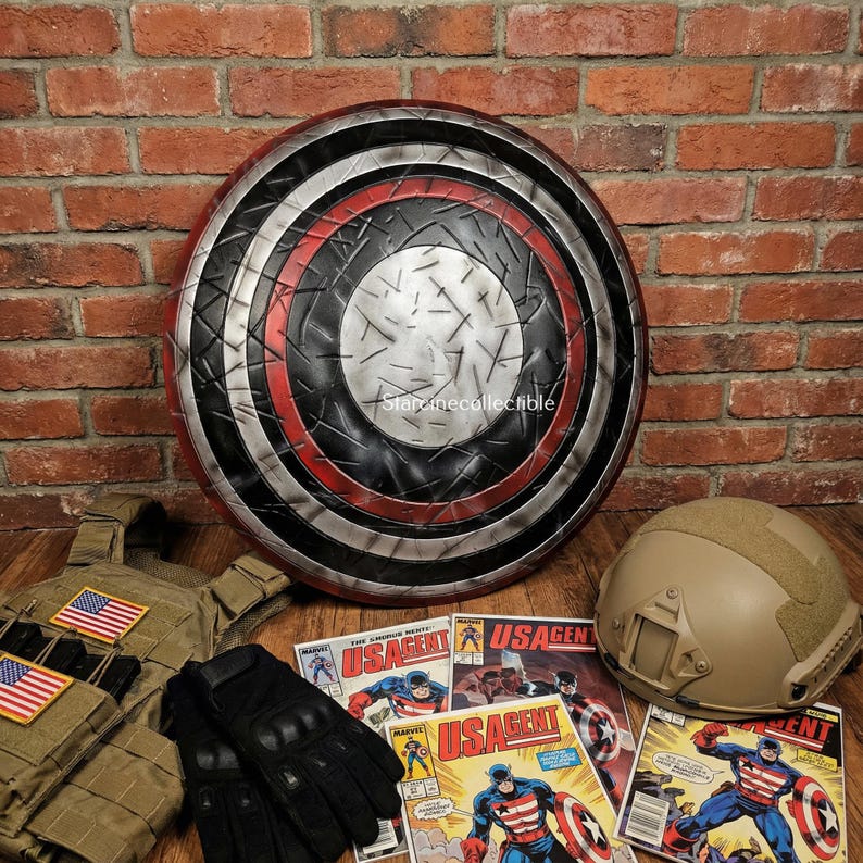 May include: A large, round shield with a red, black, and silver design. The shield is surrounded by a tactical vest, gloves, a helmet, and comic books featuring a superhero. The items are arranged on a wooden surface.