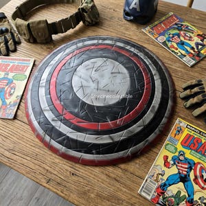 May include: A Captain America shield with a red, white, black, and silver design. The shield is circular and has a textured surface. Comic books and military gear are also visible.