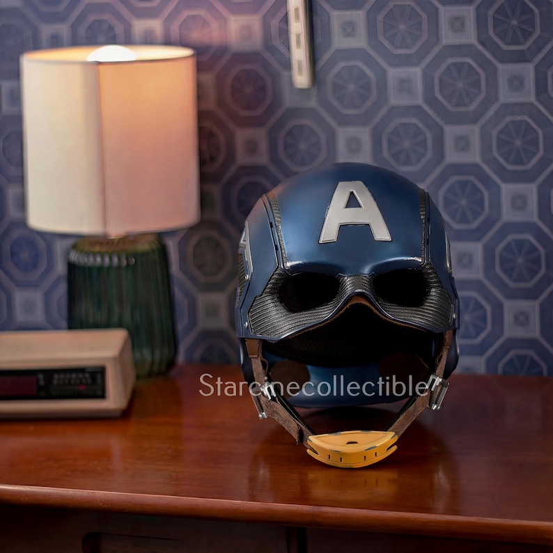 May include: A blue Captain America helmet with a silver "A" on the front. The helmet has a black face guard and a yellow chin strap. The helmet is sitting on a wooden surface.