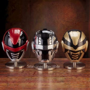 Puede incluir: Tres cascos futuristas en rojo, plateado y dorado, cada uno sobre un soporte plateado. Los cascos tienen diseños detallados de viseras y caras. Se exhiben sobre una superficie de madera con un fondo marrón oscuro. El texto "Starcinecollectible" es visible.